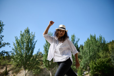 Woman dancing outdoors wearing a white hat and shirt, enjoying a sunny day with clear blue skies and...