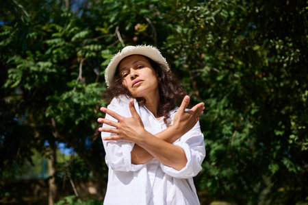 Confident woman in a white hat crossing arms in front of a lush green backdrop, basking in the...