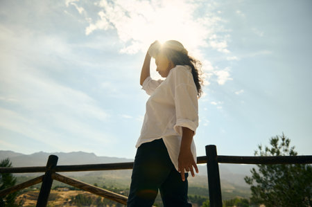 Woman in white shirt standing in the sunlight, outdoors with scenic mountains in the background,...