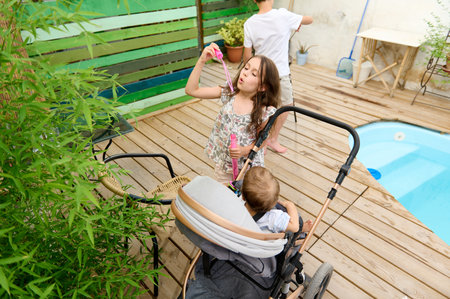 A young girl is blowing bubbles beside a baby in a stroller by the pool on a wooden deck. The scene...
