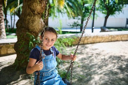 A happy young girl swings in a sunny park, smiling and enjoying her outdoor playtime. The lush green...