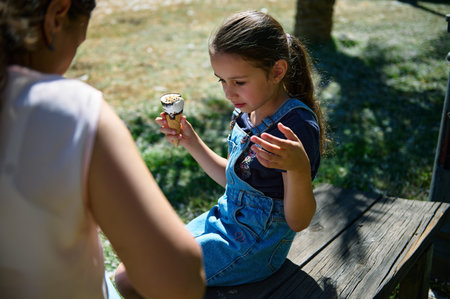 A young girl holding an ice cream cone enjoys a summer day outdoors, accompanied by another person....