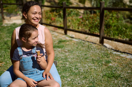Smiling mother and daughter sitting outside on a sunny day while the young girl enjoys an ice cream...