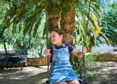 Smiling young girl swinging in the park under the shade of trees, enjoying a sunny summer day. The...