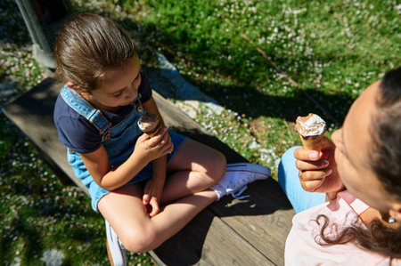 A mother and daughter sit together enjoying ice cream cones on a sunny day. They share a moment of...