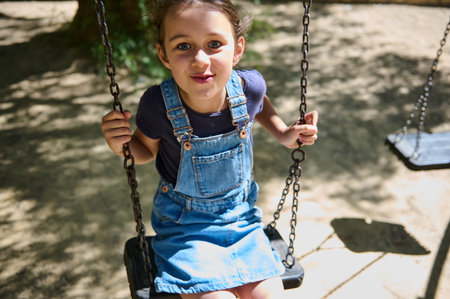 A cheerful girl in denim overalls enjoys swinging in a park on a sunny day. The bright and playful scene captures a moment of carefree childhood joy.の写真素材