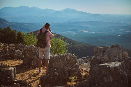A hiker with a backpack taking photos of a scenic mountain landscape from a rocky vantage point during a clear day.の写真素材