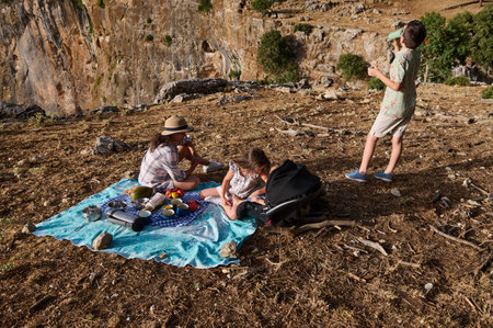 A family enjoying a picnic on a rocky hillside. The scene features a mother and children with picnic items on a blanket, surrounded by nature and mountains.の写真素材