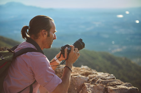 Side view of a photographer taking photos of a beautiful mountain landscape, focusing with a professional camera. The scene captures the essence of outdoor adventure and nature photography.の写真素材