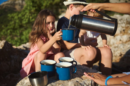 A family enjoys a camping trip in the mountains, with children being served hot beverages in blue mugs from a thermos, amid a natural, scenic environment.の写真素材