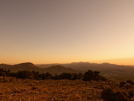 A tranquil mountain landscape at sunset, casting an orange glow over the distant hills and clear sky. The rugged terrain and dramatic lighting create a peaceful and captivating scene.の写真素材
