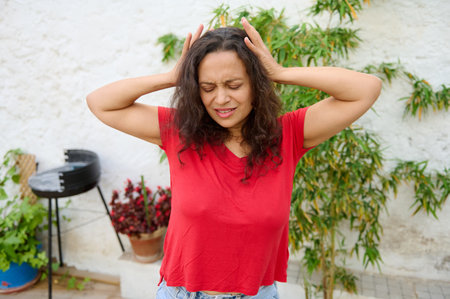 A woman in a red shirt holding her head in frustration and discomfort while standing outside. The...
