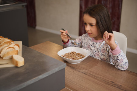 A young girl is seated at a wooden table eating a bowl of cereal for breakfast. Sliced bread is visible on the counter nearby, creating a cozy breakfast setting.の写真素材