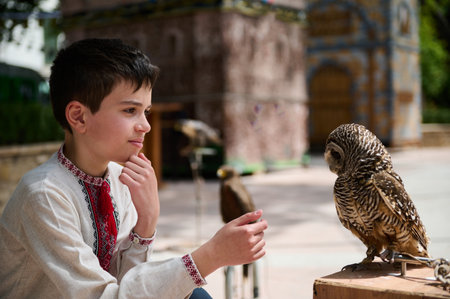Young boy in traditional attire interacts with a perched owl in an outdoor setting, showcasing...