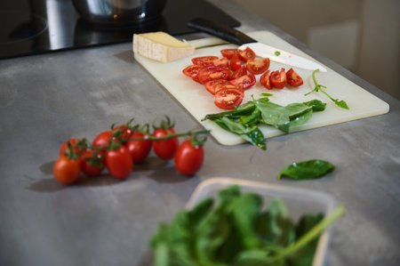 A kitchen countertop featuring freshly cut tomatoes, leafy greens, and cheese on a cutting board. Modern kitchen preparation scene highlighting fresh ingredients.の写真素材