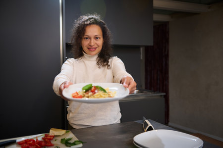 A happy woman offers a delicious plate of pasta in a contemporary kitchen setting. Fresh ingredients are visible on the counter.の写真素材