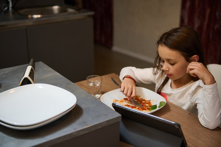 A young girl is enjoying a pasta meal while watching content on a digital device at a kitchen table. The scene showcases a cozy home environment.の写真素材
