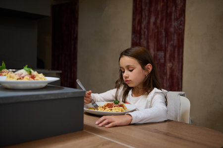 A young girl is sitting at a dining table and eating a plate of pasta. The setting appears to be indoors in a cozy environment.の写真素材