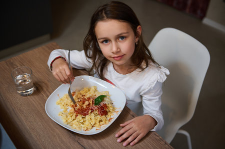 A young girl sitting at a wooden table, enjoying a plate of pasta with tomato sauce and cheese, with a glass of water beside her. She looks content and focused.の写真素材