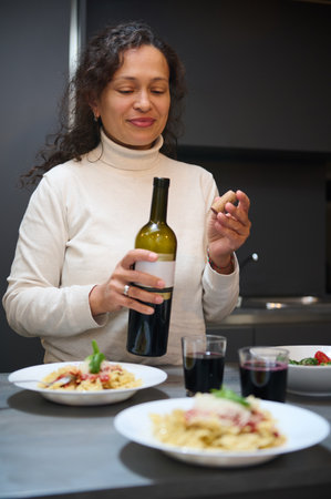 Woman opening a wine bottle in a modern kitchen with a table set with pasta dishes and glasses. Cozy...