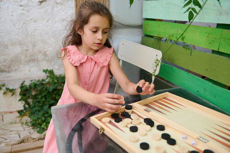 A young girl in a pink dress is focused on playing a backgammon game outside. She is arranging black...