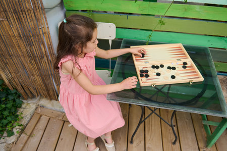 A young girl in a pink dress is playing backgammon on a glass table outside on a wooden deck. The...