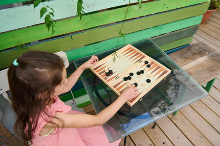 A young girl enjoys playing a game of backgammon outdoors, seated at a glass-topped table against a...