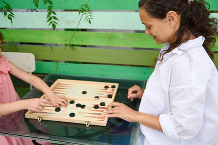 A mother and daughter enjoying an outdoor game of backgammon on a glass table. The background...