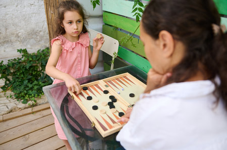 A mother and daughter bond while playing backgammon outdoors. The girl, dressed in pink, focuses...