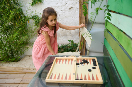 A young girl in a pink dress engaging in a game of backgammon outside in a green, garden setting,...