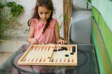 A young girl in a pink dress playing backgammon outdoors on a glass table. The setting is surrounded...