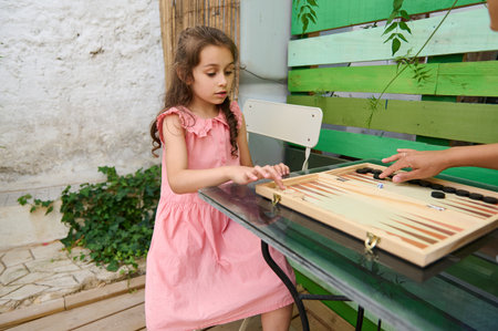 A young girl in a pink dress playing a backgammon game outdoors. The scene captures leisure,...