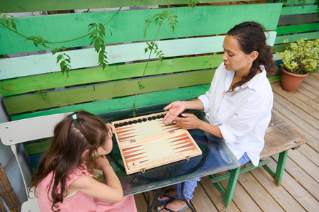 Mother and daughter playing backgammon outside on a sunny day. Family bonding and quality time spent playing a classic board game in a garden setting.の写真素材