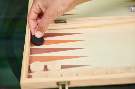 Close-up of a hand placing a black chip in a backgammon game on a wooden board. The image captures...