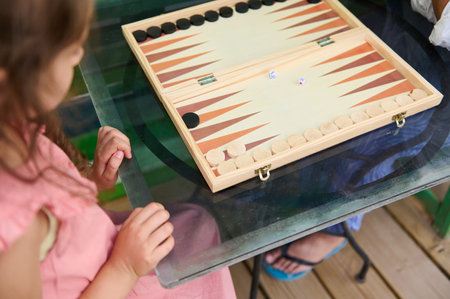 A young girl in a pink dress playing backgammon on a wooden board with dice. Indoor setting,...