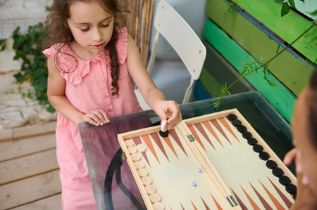 A young girl in a pink dress is playing a board game outdoors on a patio. The scene is set on a...
