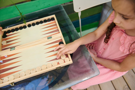 A young girl in a pink dress plays backgammon on a glass table outdoors. The image captures a moment...