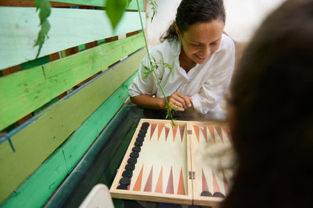 A smiling woman playing backgammon with a friend in an outdoor setting. The scene captures leisure,...