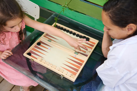 A young mother and her daughter bonding while playing a game of backgammon outdoors on a glass...