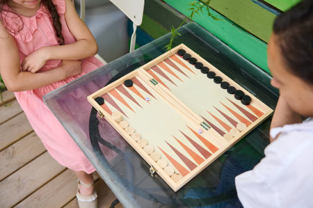 Two children are enjoying a game of backgammon on a glass table outdoors. The friendly atmosphere...