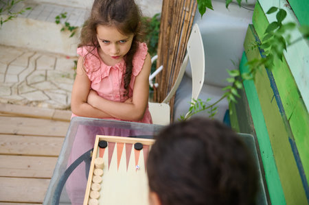 A young girl in a pink dress intensely playing backgammon with a friend outdoors. Her serious...