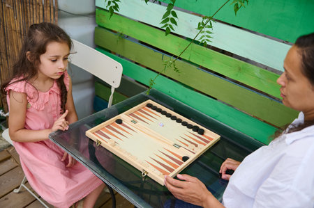 Mother and daughter enjoy a game of backgammon together outdoors. The setting is bright with a green...