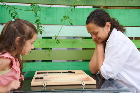 A loving mother and her young daughter share quality time playing backgammon together in a serene...