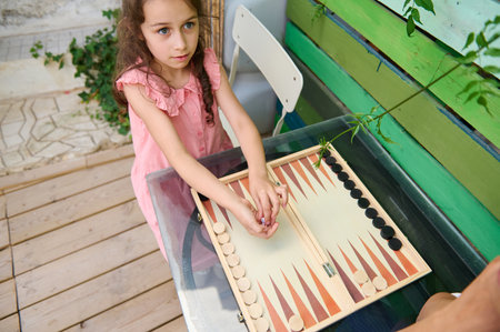 Young girl in a pink dress playing backgammon outdoors at a wooden table with a colorful background....