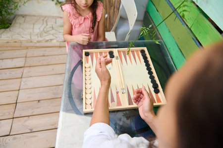 A mother and her daughter engage in a fun game of backgammon at an outdoor table. The family bonding...