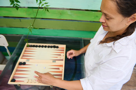 A woman in a white shirt engaged in a game of backgammon outdoors. The table features a vibrant...
