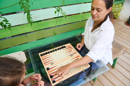 Mother and daughter enjoying a game of backgammon outdoors, fostering family bonding. The image...