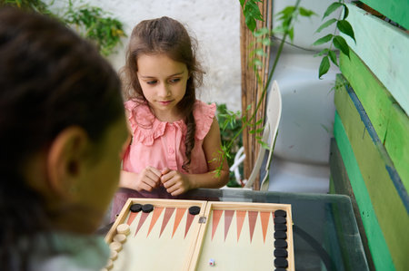 Two young girls engrossed in a game of backgammon while sitting outdoors. The children are...