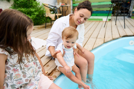 A mother spending quality time with her children by the poolside. The baby is being held while another child sits close by, all enjoying the sunny day and the refreshing water.の写真素材