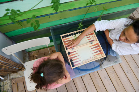 Top view of two people enjoying a game of backgammon on a glass table outdoors. The vibrant green...